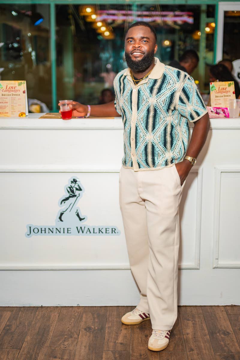 Ricardo Casanova, junior channel development manager at Red Stripe, leaning into the Johnnie Walker bar in a patterned knit short-sleeve shirt, cream trousers, and classic low tops.