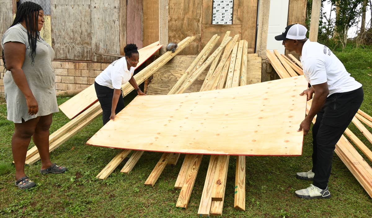 From left: Tanekia Titus looks on as Hermine Pryce Foundation co‑founders Andrea Pryce and O’Neil Pryce lift a sheet of plywood donated to her at her home in Bunkers Hill, Trelawny.
