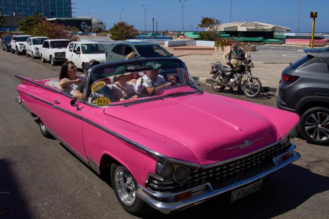 Tourists travel in a classic American car next to a line of drivers waiting to buy fuel for their cars in Havana, Cuba, on February 16, 2026. 