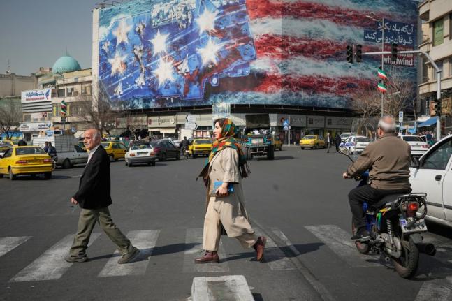 Pedestrians walk past a billboard depicting a US aircraft carrier with damaged fighter jets on its deck and a sign in Farsi and English reading, ‘If you sow the wind, you’ll reap the whirlwind,’ at Enqelab-e-Eslami (Islamic Revolution) Square in Tehr