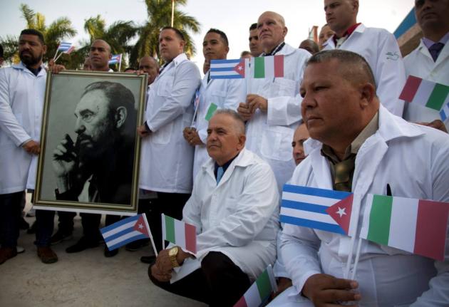 In this March 2020 photo, Cuban doctors and medical professionals pose for the media with a photo of Fidel Castro and flags of Italy and Cuba, prior to their departure to Italy to assist with the COVID-19 pandemic. 